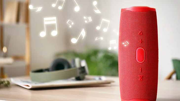 Na stole stoi czerwony głośnik. W tle widać słuchawki i nuty muzyczne.