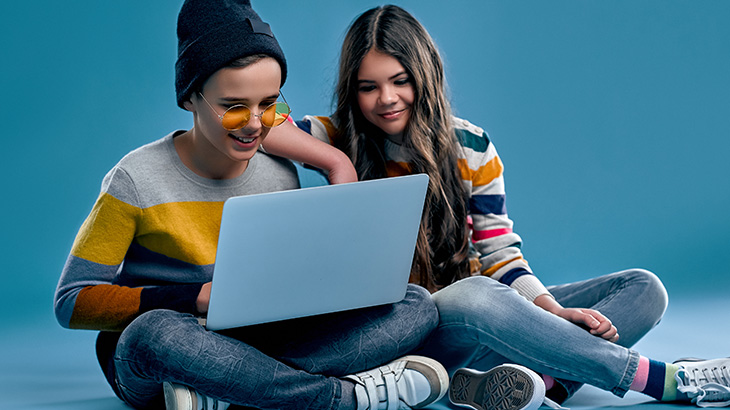 Dwoje nastolatków siedzi na podłodze, patrząc na laptopa. Chłopak ma czapkę i okulary, a dziewczyna w paski sweter. Tło jest niebieskie.