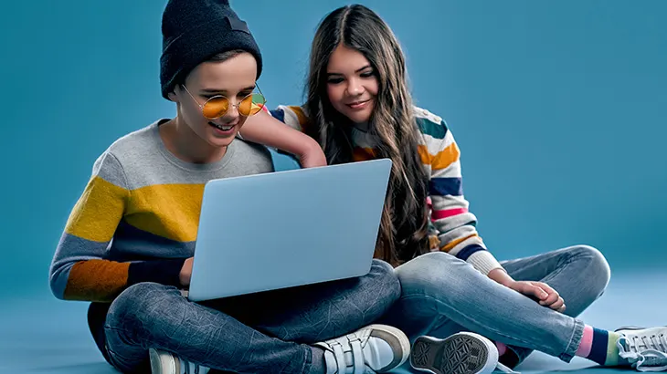 Dwoje nastolatków siedzi na podłodze, patrząc na laptopa. Chłopak ma czapkę i okulary, a dziewczyna w paski sweter. Tło jest niebieskie.