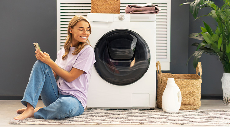 Kobieta siedzi na dywanie z telefonem przy pralce. Obok kosz i roślina doniczkowa. W tle ściana i żaluzje.
