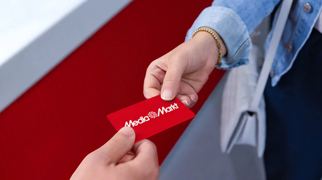 Dwie dłonie wymieniają się czerwoną kartą Media Markt na tle czerwono-białym. Widoczna część osoby z torbą i kurtką.