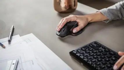 Na biurku leżą dokumenty, długopis, ołówek, klawiatura i mysz, którą trzyma dłoń. W tle widać metalowy wazon.