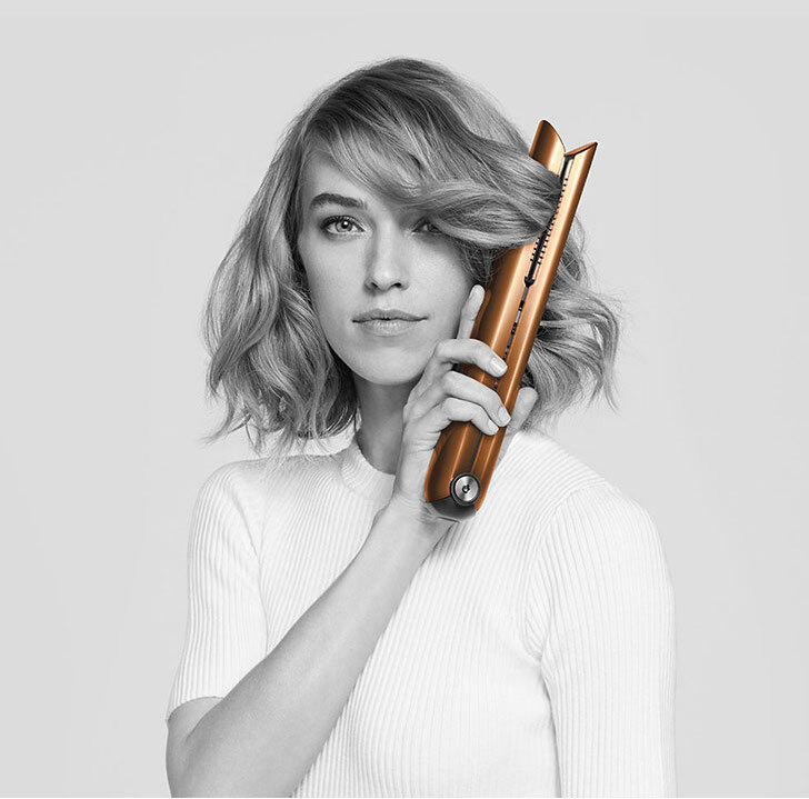 Kobieta trzyma prostownicę do włosów koloru miedzianego. Ubrana jest w białą koszulkę na tle szarego tła.