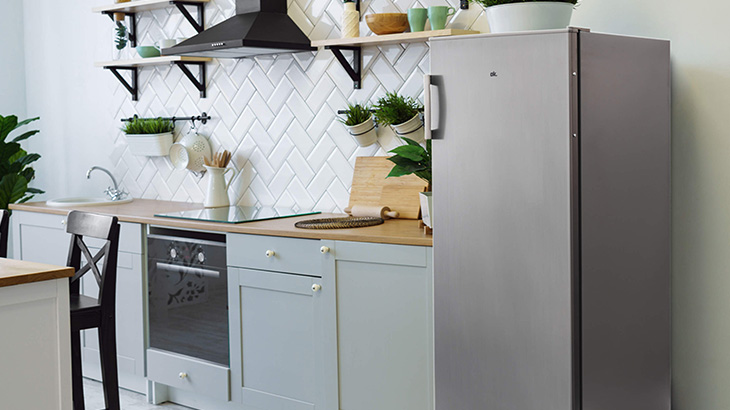 Kuchnia z szarą lodówką, białymi płytkami ułożonymi w jodełkę i drewnianymi blatami. Na ścianach półki z przyborami.