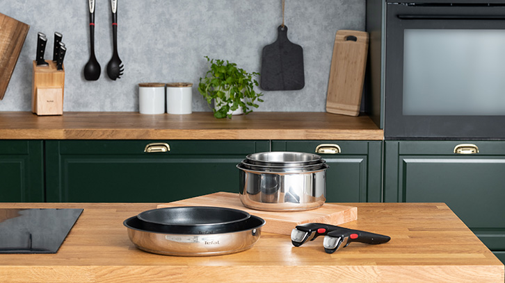 Kuchnia z drewnianym blatem, zielonymi szafkami i srebrnymi garnkami. Na blacie płyta indukcyjna, patelnia i uchwyt.