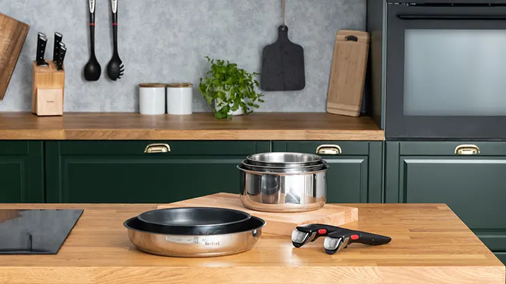 Kuchnia z drewnianym blatem, zielonymi szafkami i srebrnymi garnkami. Na blacie płyta indukcyjna, patelnia i uchwyt.