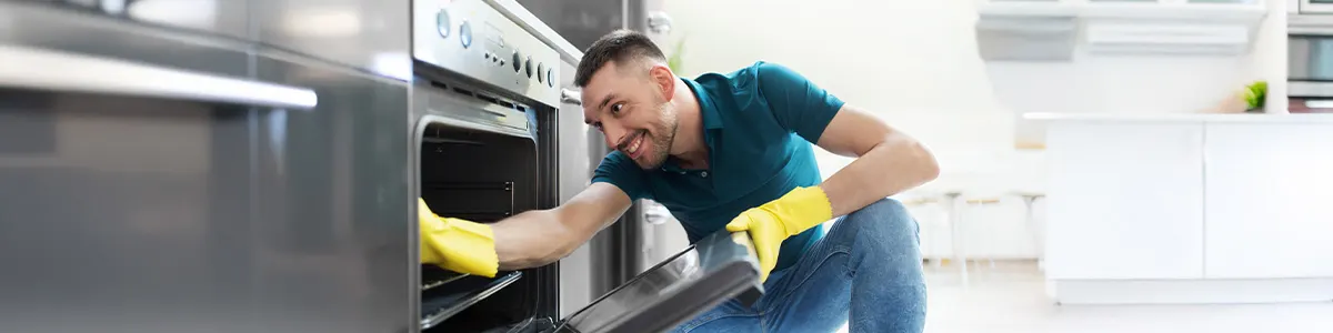 Osoba w zielonych rękawiczkach czyści piekarnik niebieską szmatką. Piekarnik ma srebrny panel sterowania z pokrętłami.