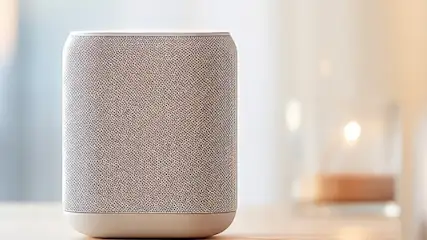 Na pierwszym planie stoi szary głośnik na stole. W tle widać kanapę z białym kocem i rozproszone światło.