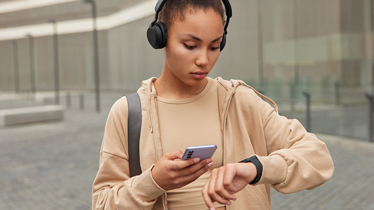 Kobieta w słuchawkach sprawdza telefon i zegarek, w tle budynek.