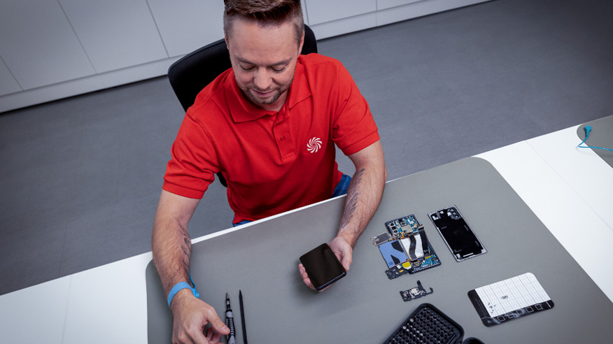 Mężczyzna w czerwonej koszulce polo siedzi przy stole z rozłożonym telefonem i narzędziami do naprawy.
