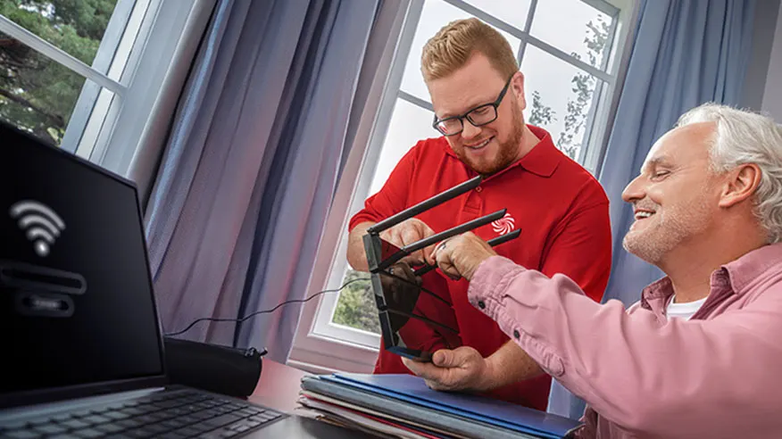 Dwóch mężczyzn konfiguruje router Wi-Fi przy biurku z laptopem, na tle okna z zasłonami.