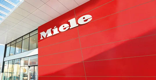 Czerwona ściana budynku z białym logo "Miele" i przeszklone okna po lewej stronie.