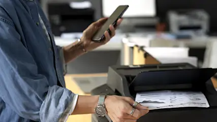 Osoba trzyma telefon i wyjmuje wydrukowaną kartkę z drukarki. W tle biuro z urządzeniami.