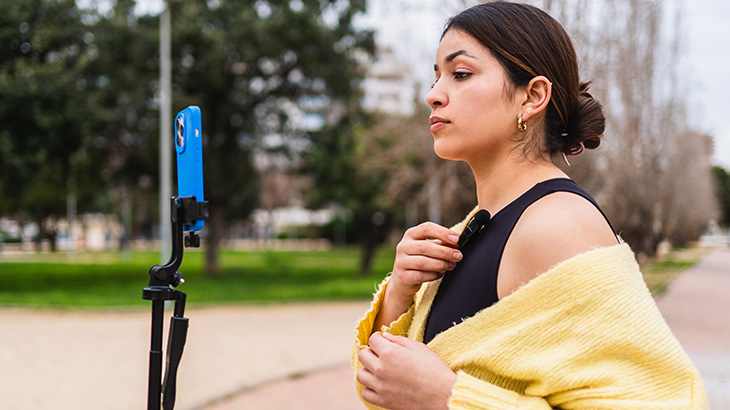 Kobieta w parku używa smartfona umieszczonego na statywie, aby nagrać wideo, patrząc w ekran urządzenia, poprawia bezprzewodowy mikrofon przypięty do bluzki.