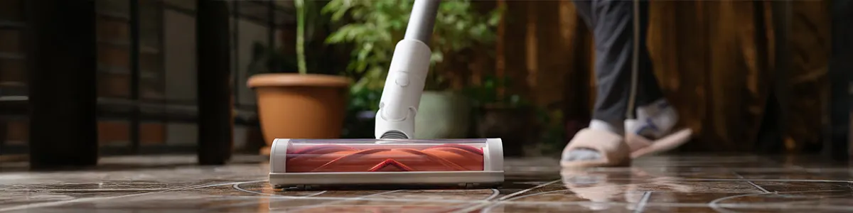 Osoba odkurza brązową podłogę odkurzaczem z pomarańczową szczotką. W tle doniczka z zieloną rośliną i ciemne meble.