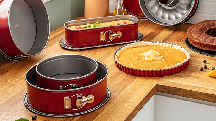 Na drewnianym blacie stoją czerwone formy do pieczenia, tarta cytrynowa, babka i inne ciasta.