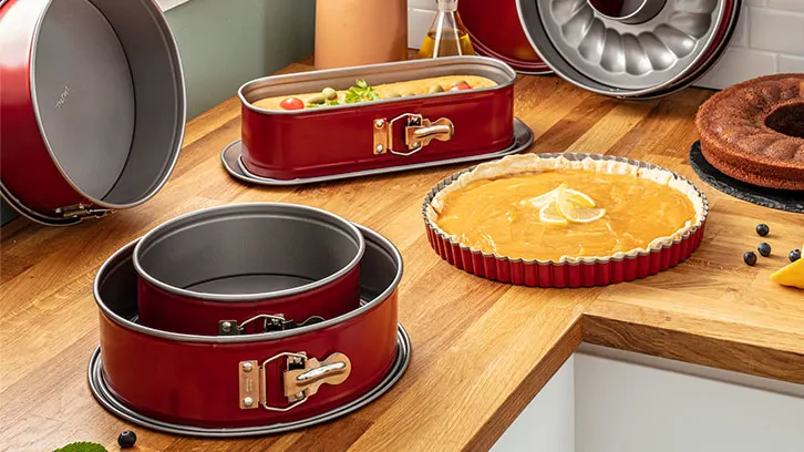 Na drewnianym blacie stoją czerwone formy do pieczenia, tarta cytrynowa, babka i inne ciasta.