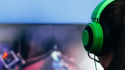 Osoba w zielonych słuchawkach gra w grę wideo na monitorze na tle niebieskiej ściany.