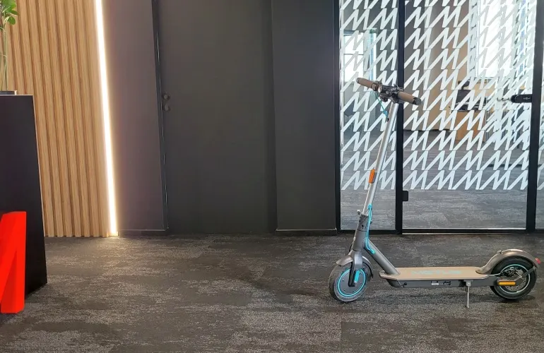 Na szarym dywanie stoi szara hulajnoga elektryczna, tło to ściana i szklane drzwi z białym wzorem.