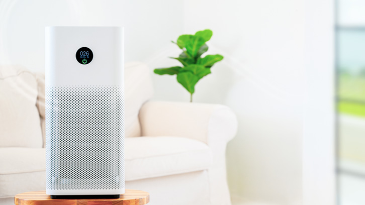 Biały oczyszczacz powietrza stoi na drewnianym stoliku, z tyłu biały fotel i zielona roślina na białym tle ściany.