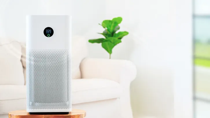 Biały oczyszczacz powietrza stoi na drewnianym stoliku, z tyłu biały fotel i zielona roślina na białym tle ściany.