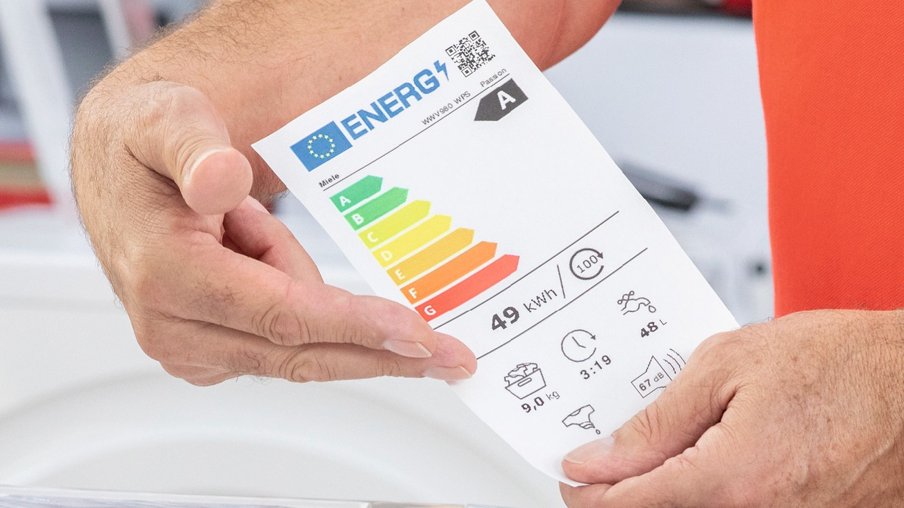 Mężczyzna trzyma etykietę energetyczną z klasą A, zużyciem 49 kWh, wagą 9 kg, czasem prania 3:19, zużyciem wody 48L i hałasem 67 dB.