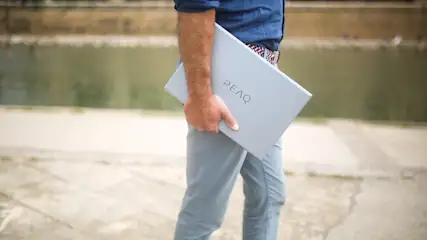 Osoba trzyma szary laptop marki PEAQ na tle wody i betonowej nawierzchni.