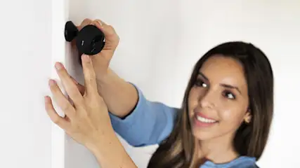 Kobieta w niebieskiej bluzce montuje czarną kamerę na białej ścianie.