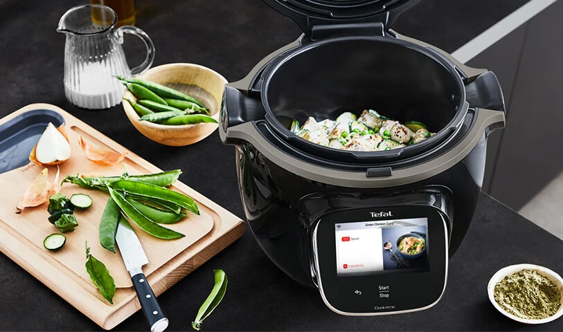 Czarny szybkowar elektryczny Tefal z wyświetlaczem, obok deska z nożem, groszek, cebula i dzbanek.