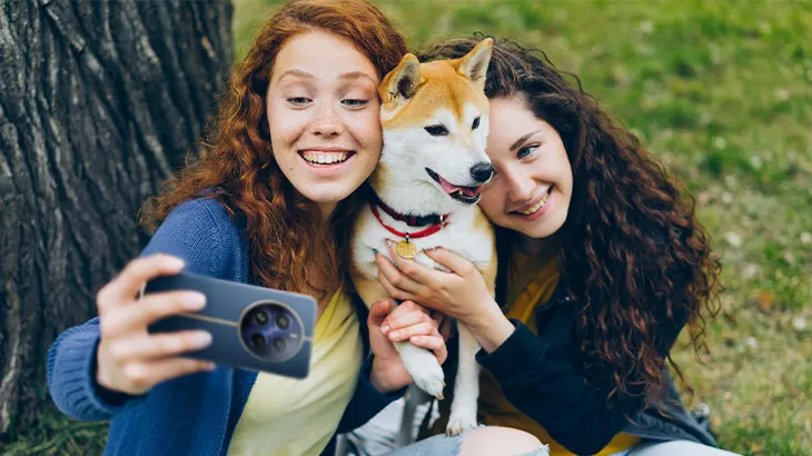 Dwie kobiety robią sobie selfie z psem rasy Shiba Inu na tle drzewa i trawy.