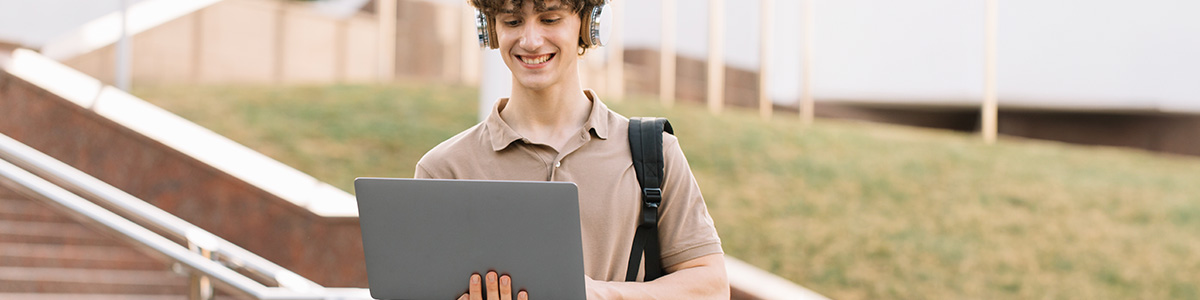Na tle schodów osoba korzysta z laptopa, ma na sobie słuchawki i ramię od plecaka
