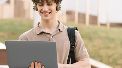 Na tle schodów osoba korzysta z laptopa, ma na sobie słuchawki i ramię od plecaka