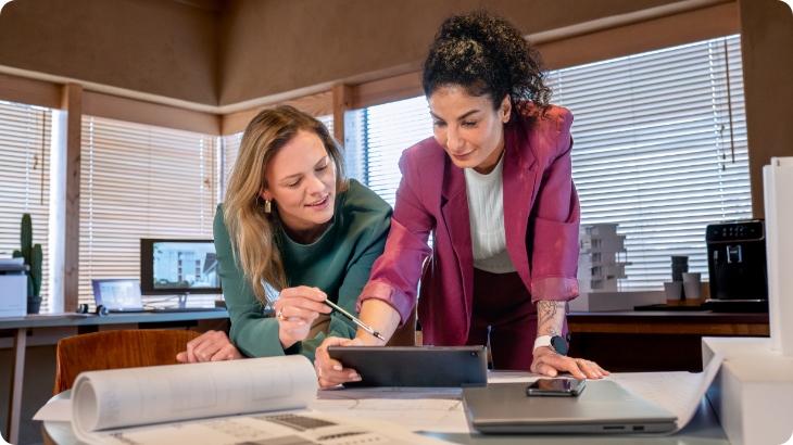 Dwie kobiety w biurze patrzą na tablet. Na stole leżą dokumenty, laptop i telefon. W tle okno z żaluzjami.