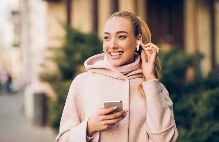 Kobieta w różowym płaszczu z kapturem trzyma telefon i wkłada słuchawkę do ucha na tle zabudowań i zieleni.