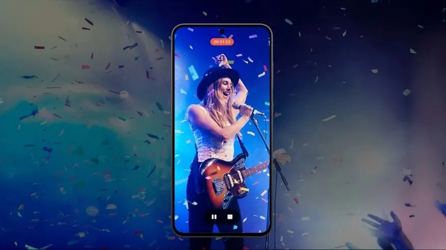 Na telefonie kobieta w kapeluszu na scenie. Obraz wychodzi poza ramki, wszędzie confetti.