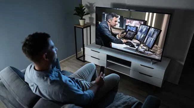 Mężczyzna siedzi na kanapie i ogląda telewizor. Na ekranie widać mężczyznę pracującego przy wielu monitorach. Obok stoi roślina doniczkowa.