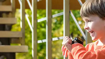 Chłopiec uśmiecha się na tle schodów z jasnego drewna z metalową barierką i zielenią w tle, trzymając w rękach czarny przedmiot.