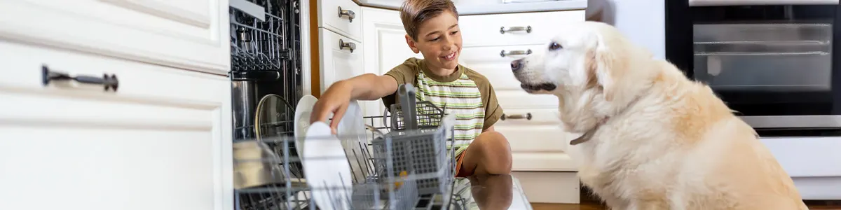 Chłopiec układa naczynia i sztućce w zmywarce w towarzystwie psa.