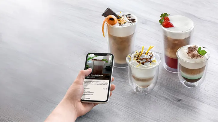 Na stole stoi telefon z przepisem na napój obok czterech szklanek z napojami warstwowymi, z bitą śmietaną i dekoracjami.