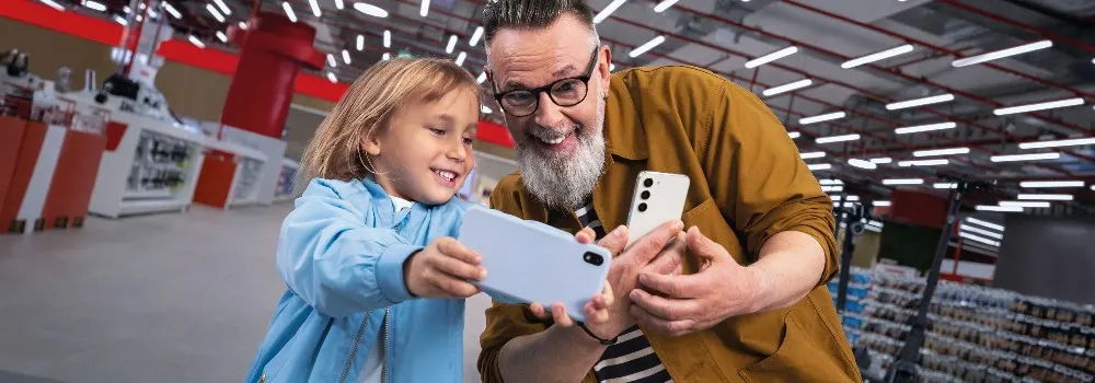 Wnętrze sklepu: mężczyzna i dziecko trzymają telefony. Mężczyzna ma brodę i okulary, ubrany w brązową kurtkę. Dziecko ma niebieską bluzkę.