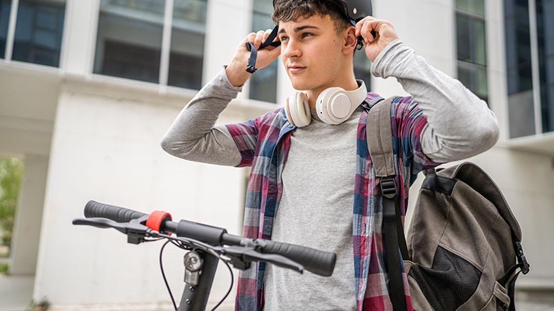 Młody mężczyzna zakłada kask obok hulajnogi, ma plecak i słuchawki na szyi. W tle budynek z oknami.