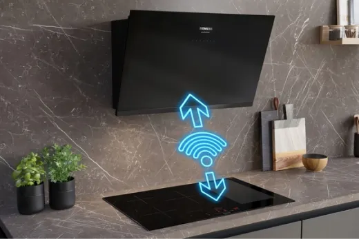 Czarny okap nad czarną płytą indukcyjną na tle ściany z marmuru. Obok deski do krojenia i rośliny w doniczkach.