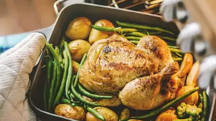 Brytfanna z pieczonym kurczakiem, ziemniakami i zieloną fasolką wyjmowana z piekarnika za pomocą rękawicy kuchennej.