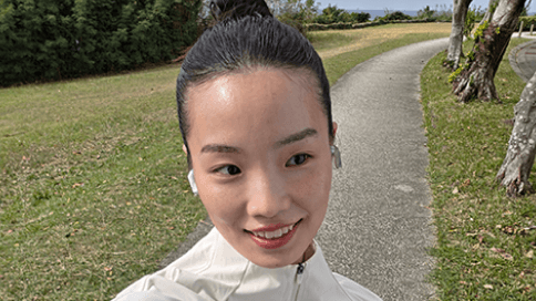 Selfie na zewnątrz: osoba w jasnej kurtce z upiętymi włosami, twarz zamazana; w tle kręta asfaltowa alejka przez zielony park z drzewami i trawnikiem.