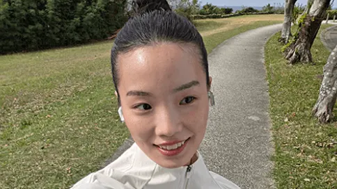 Selfie na zewnątrz: osoba w jasnej kurtce z upiętymi włosami, twarz zamazana; w tle kręta asfaltowa alejka przez zielony park z drzewami i trawnikiem.