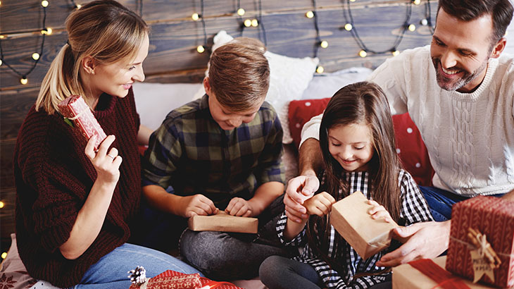 Na obrazku przedstawiona jest 4 osobowa rodzina która otwiera prezenty świąteczne.
