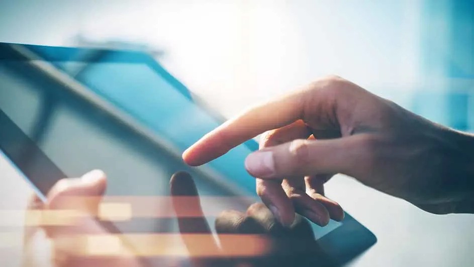 Dłoń dotyka ekranu tabletu na jasnym tle. Widoczne odbicie palca na ekranie. Urządzenie w kolorze niebiesko-szarym.