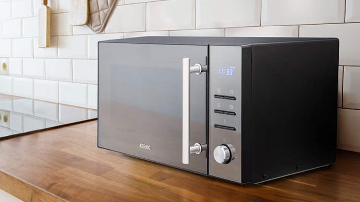 Na drewnianym blacie kuchennym stoi czarno-srebrna mikrofalówka z pionowym uchwytem i panelem sterowania po prawej stronie. W tle białe kafelki.
