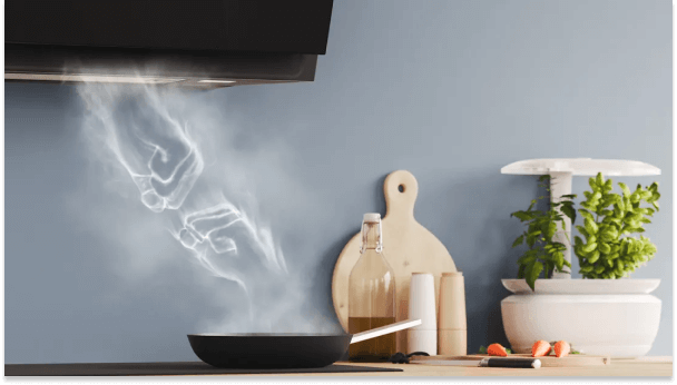Czarny okap kuchenny nad patelnią, z której unosi się para. Obok deska, butelka, przyprawy i doniczka z ziołami na tle szarej ściany.