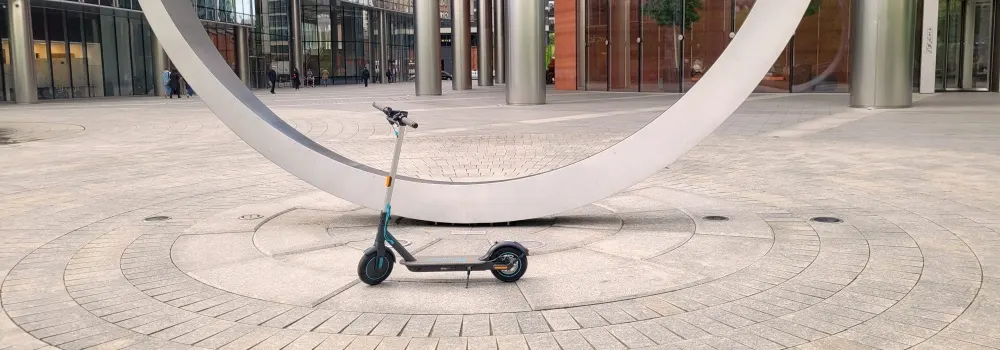 Na placu stoi hulajnoga elektryczna. W tle budynki z kolumnami i biała, łukowa rzeźba. Nawierzchnia placu z kostki, ułożona w okręgi.